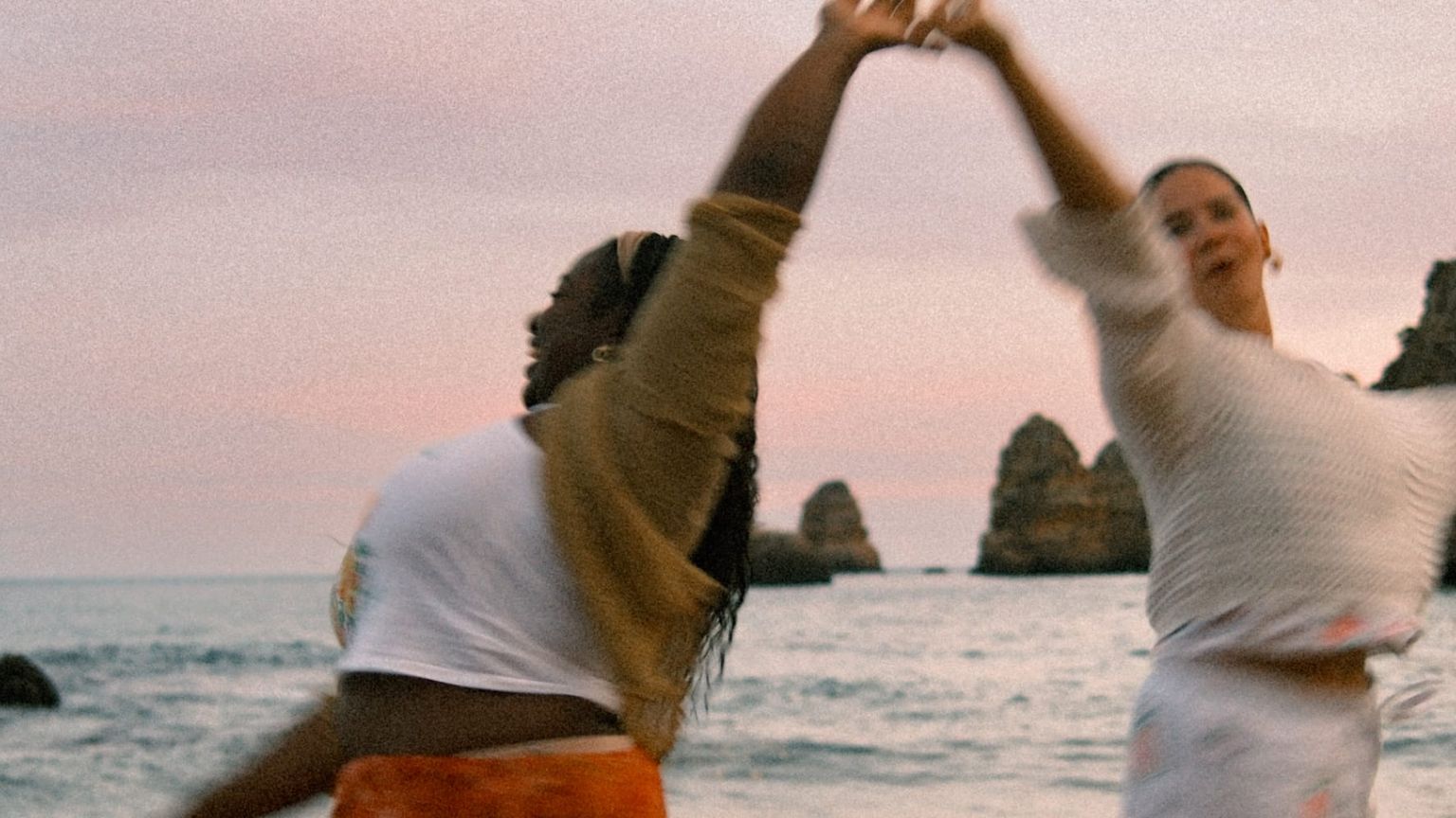 Two people joyfully dancing by the sea at sunset, surrounded by rocky cliffs and gentle waves in the background.