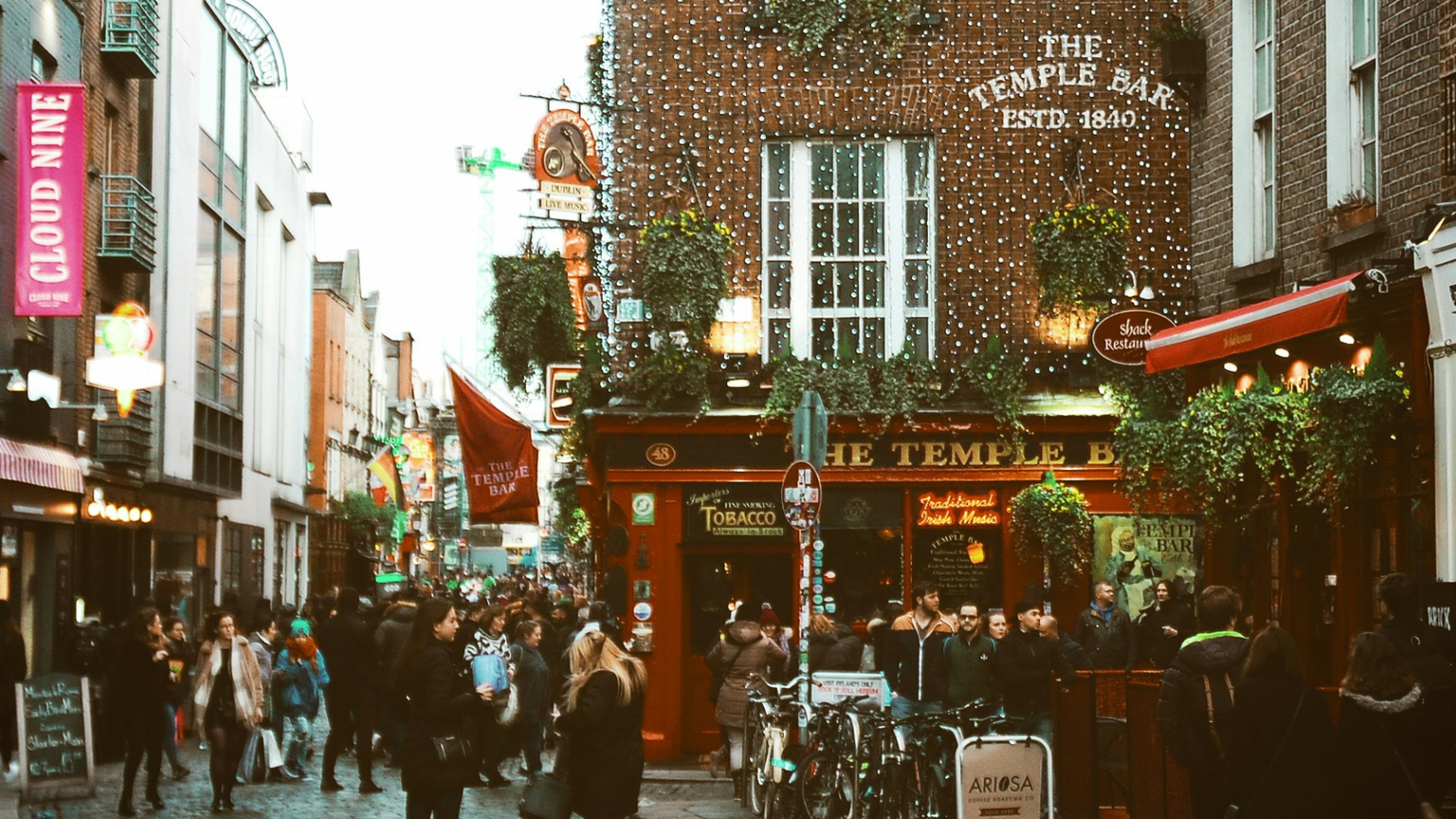 A bustling street scene at The Temple Bar in Dublin, with people walking and bicycles parked, surrounded by vibrant shops and pubs.
