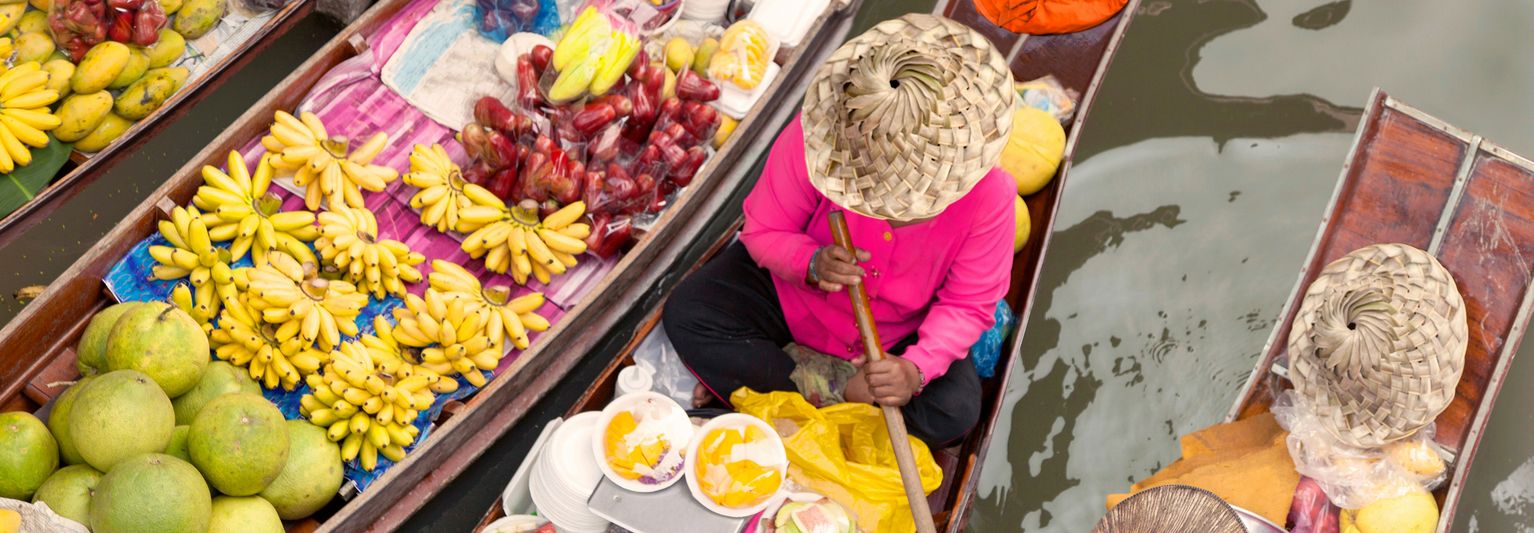 Aerial view of a person in a straw hat on a boat filled with bananas and fruit at a floating market, with water visible nearby.