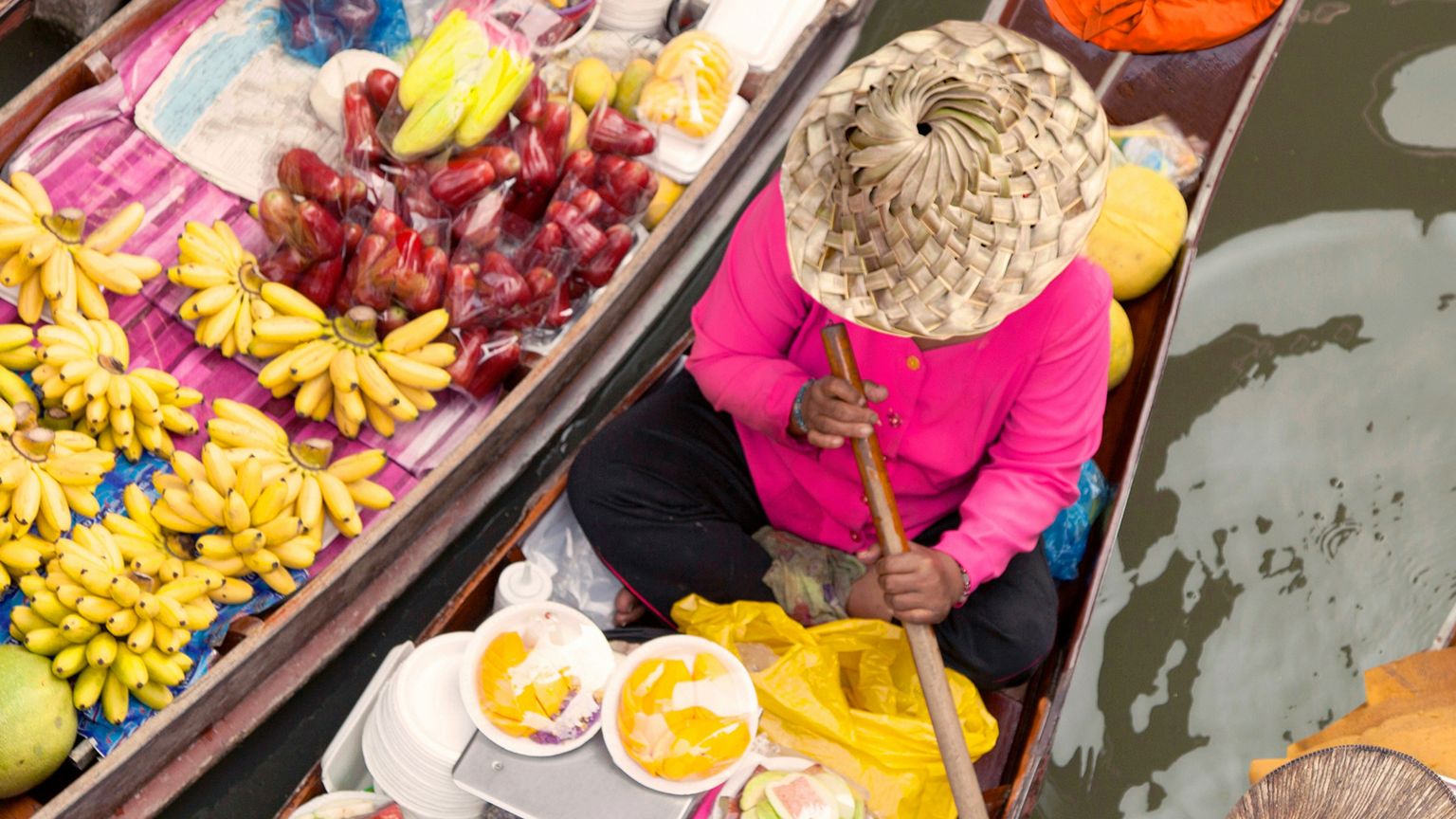 Aerial view of a person in a straw hat on a boat filled with bananas and fruit at a floating market, with water visible nearby.