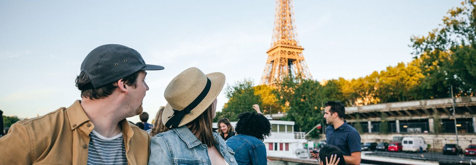 People on a boat tour in Paris, gazing at the Eiffel Tower under a clear sky, with trees and a bridge in the background.