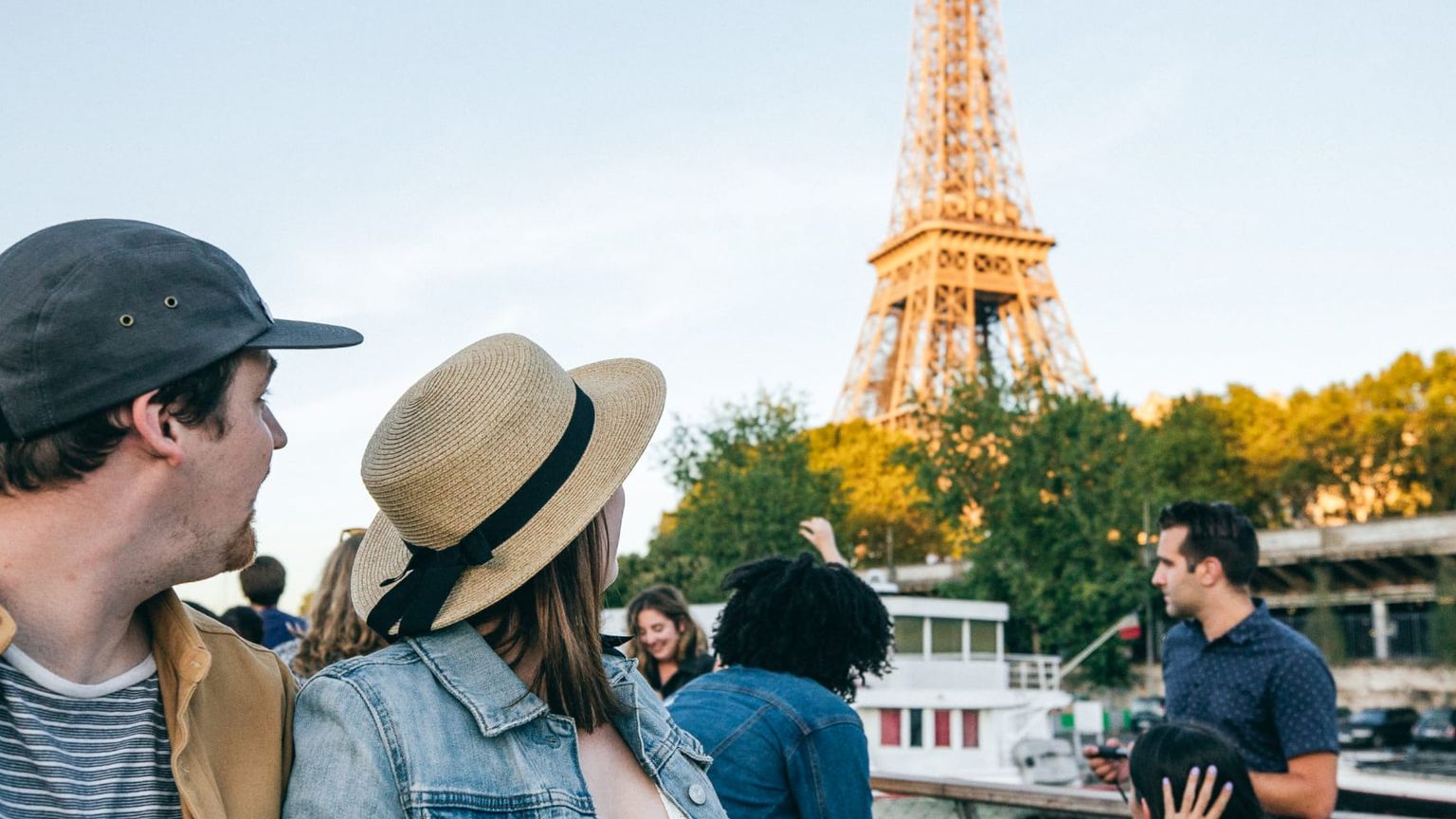 People on a boat tour in Paris, gazing at the Eiffel Tower under a clear sky, with trees and a bridge in the background.