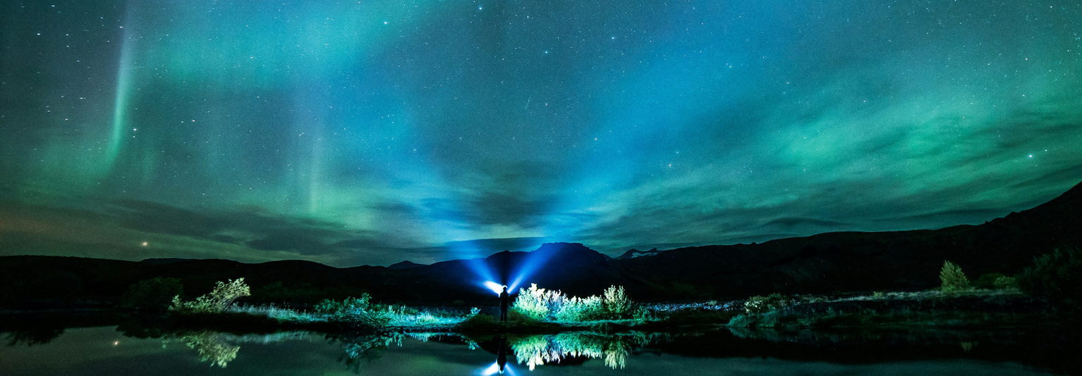 A person with a flashlight stands by a reflective lake under a vibrant green and blue aurora borealis against a starry night sky.