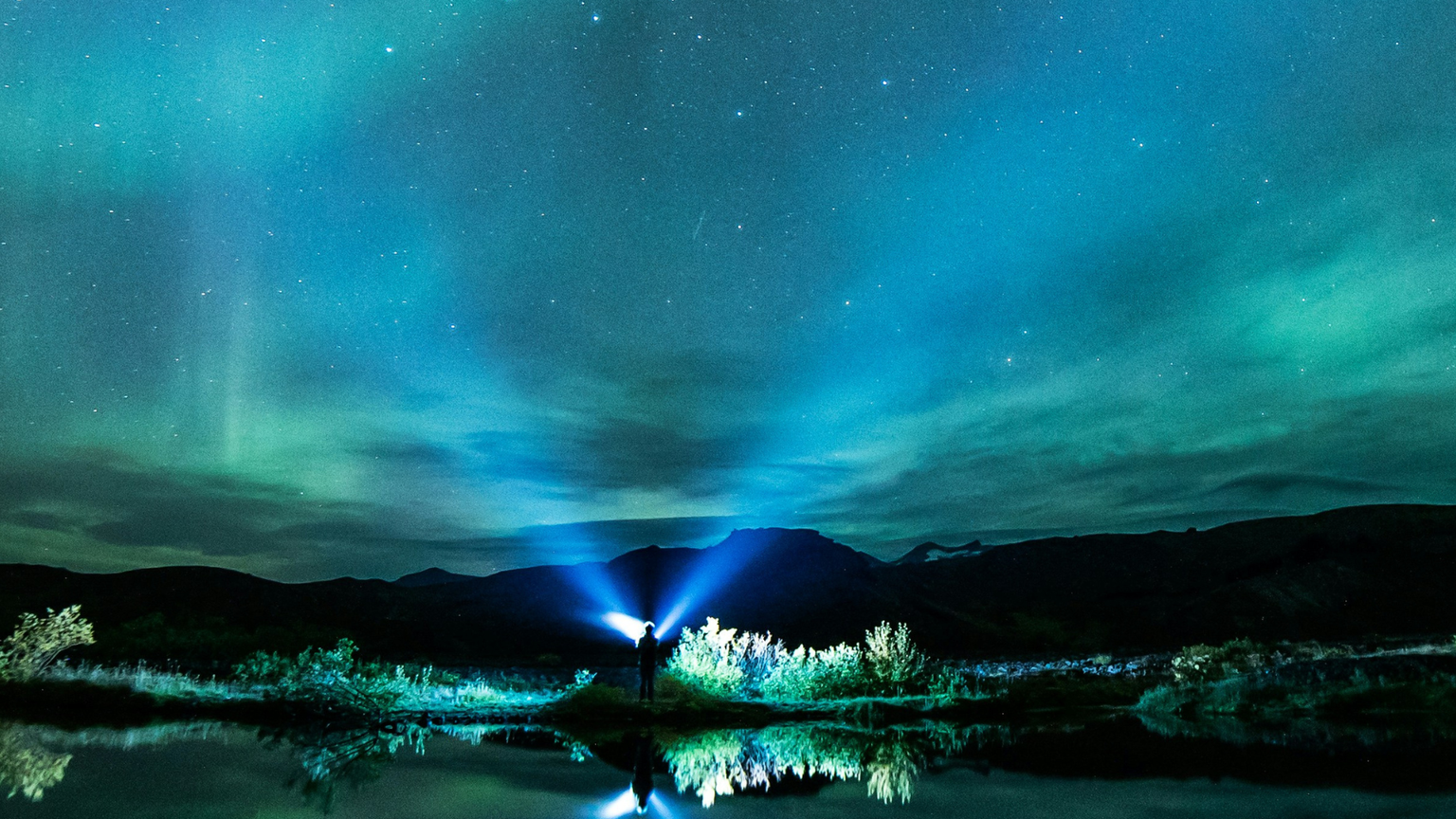 A person with a flashlight stands by a reflective lake under a vibrant green and blue aurora borealis against a starry night sky.