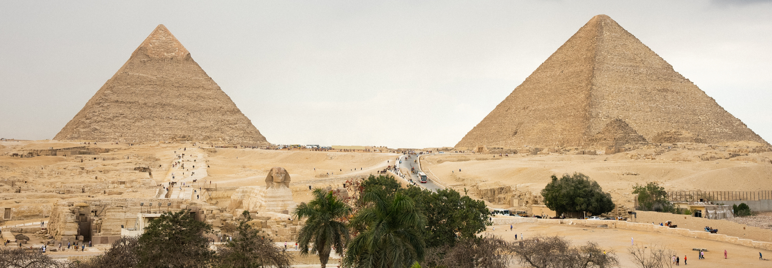 Two large pyramids in a desert landscape, with a road and scattered trees in the foreground under a cloudy sky.