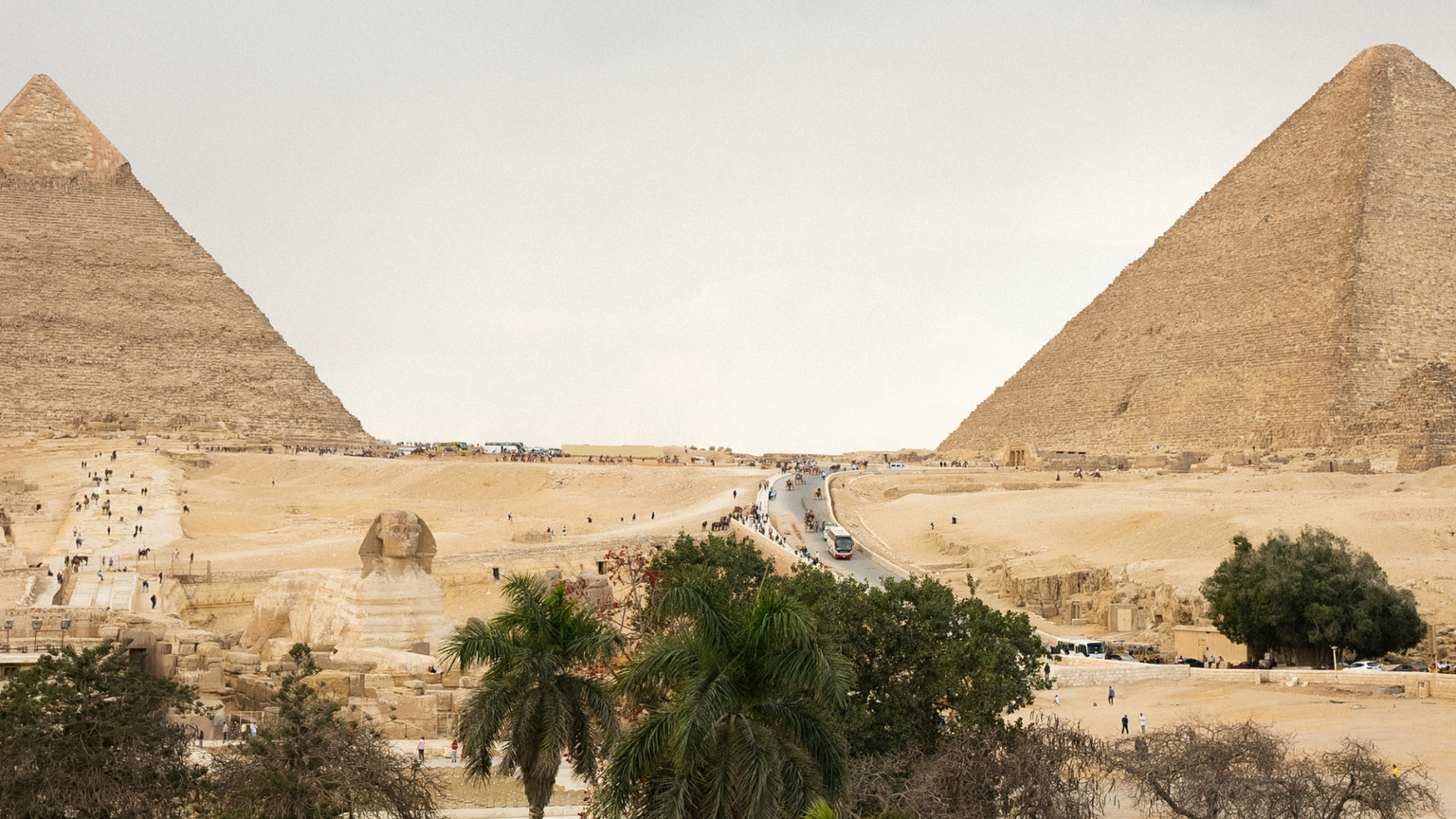 Two large pyramids in a desert landscape, with a road and scattered trees in the foreground under a cloudy sky.