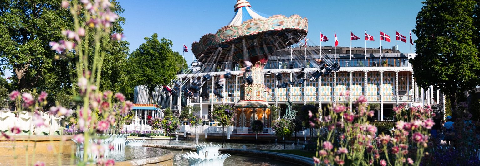 Colorful amusement park with a spinning swing ride, surrounded by lush greenery and floral gardens under a clear blue sky. Danish flags are visible.