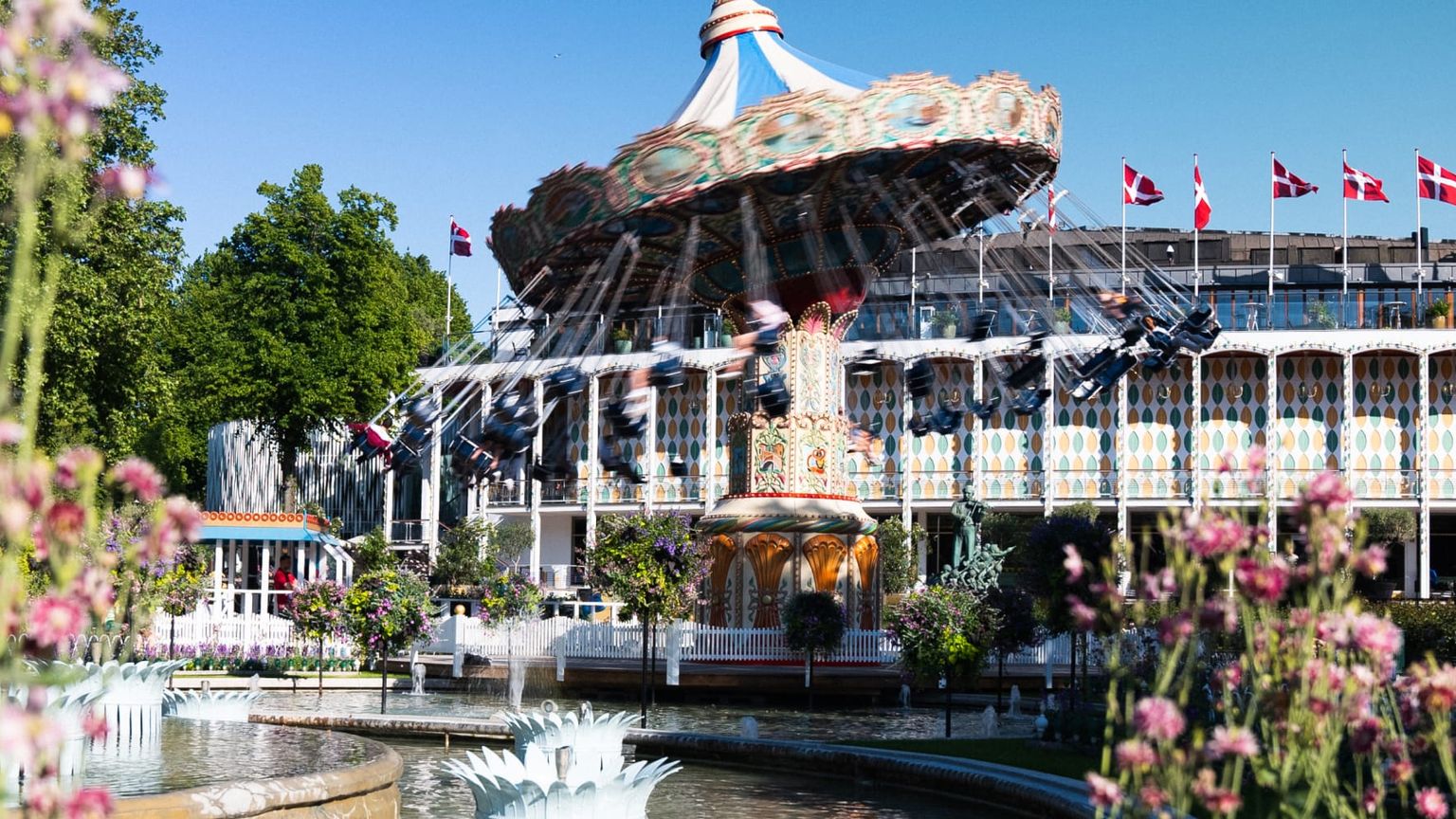 Colorful amusement park with a spinning swing ride, surrounded by lush greenery and floral gardens under a clear blue sky. Danish flags are visible.
