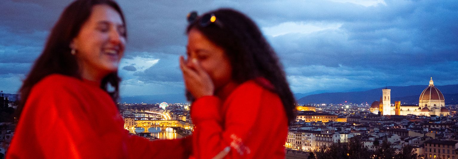 Two people laughing, wearing red jackets, with a nighttime view of a city featuring a prominent cathedral dome in the background.