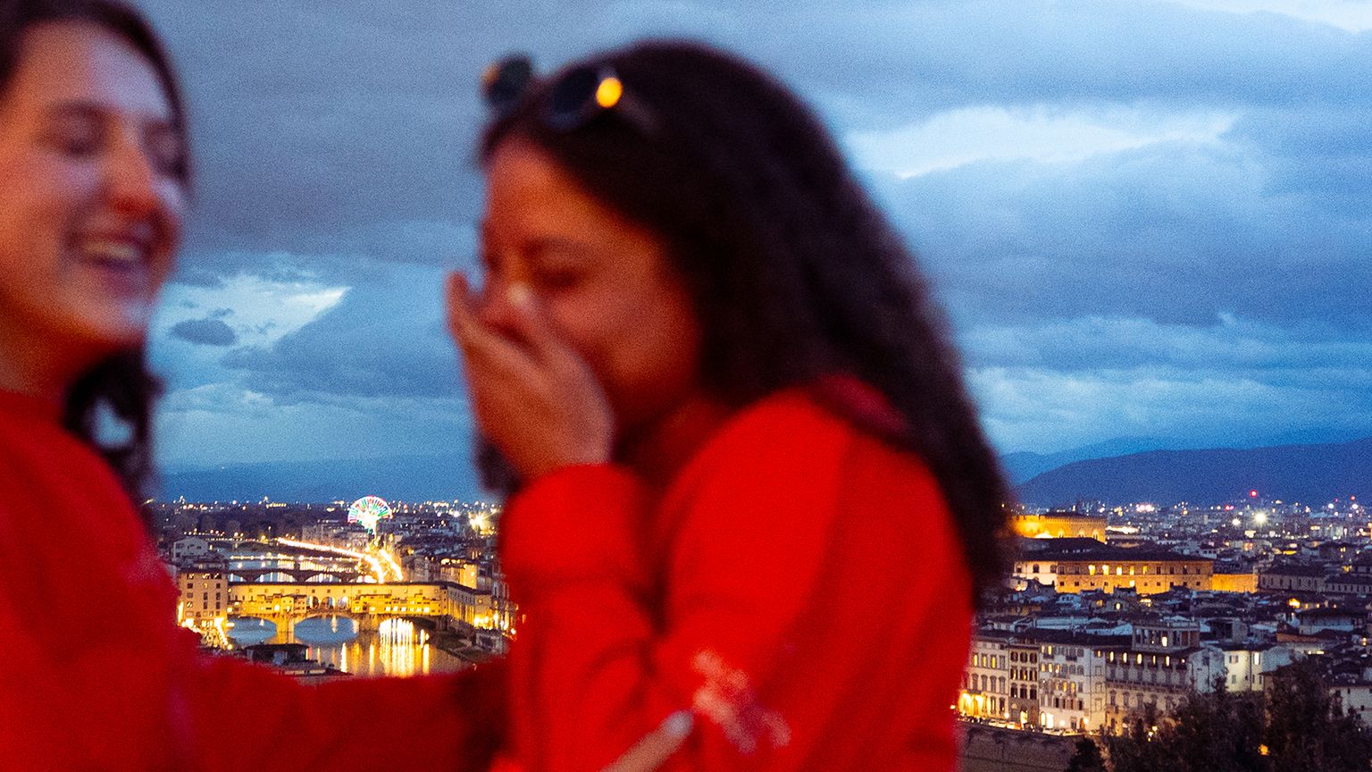 Two people laughing, wearing red jackets, with a nighttime view of a city featuring a prominent cathedral dome in the background.