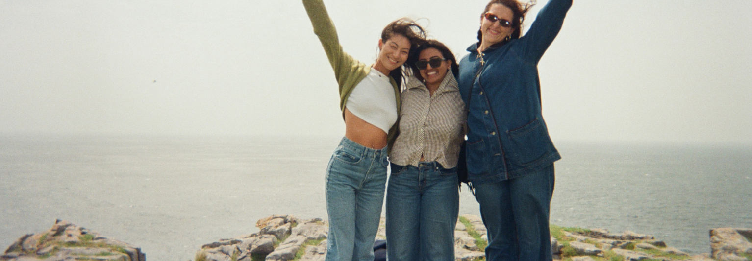 Three people smiling with arms raised, standing on a rocky cliff overlooking the ocean on a windy day.