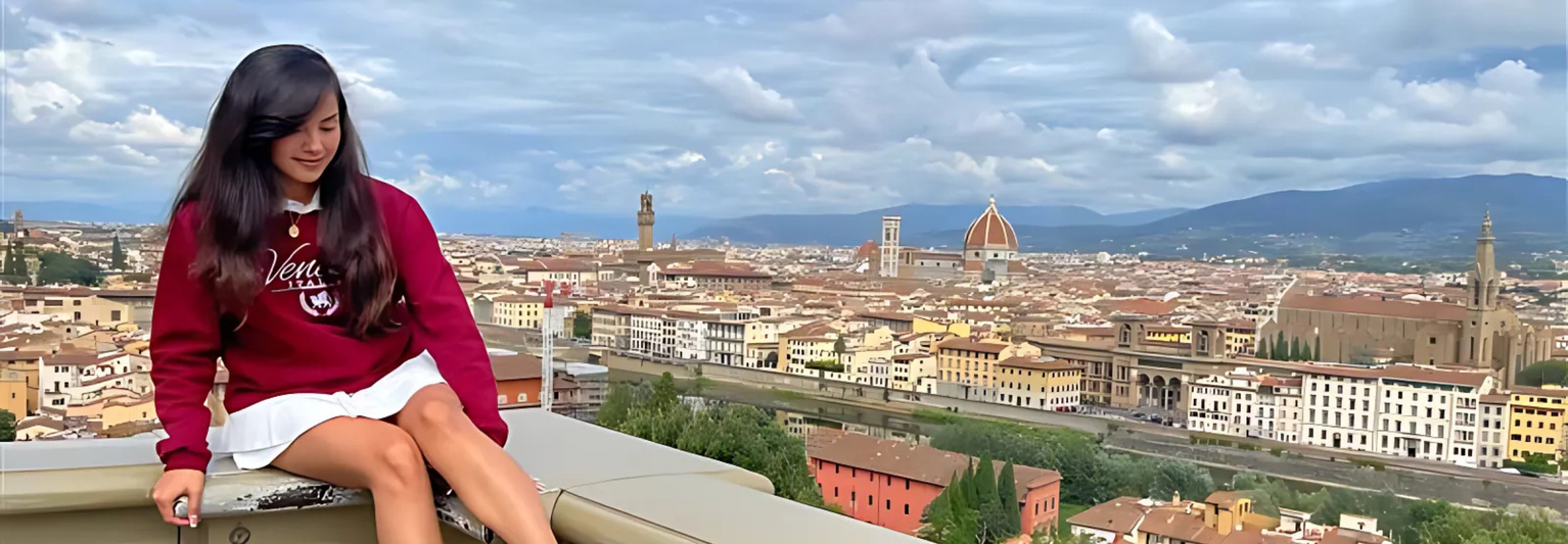A woman in a red sweatshirt sits on a ledge overlooking a cityscape with the Florence Cathedral and bell tower under a cloudy sky.