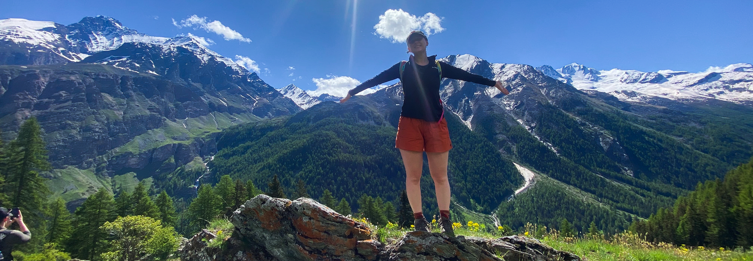 A person stands on a rock in front of snow-capped mountains and blue sky, arms outstretched. Greenery and trees surround the scene.