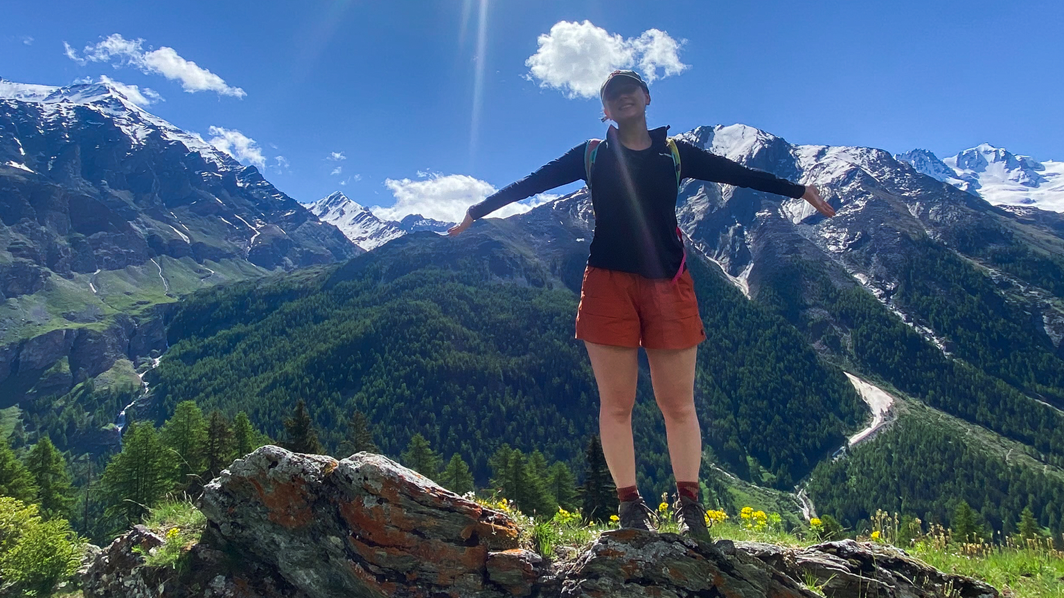A person stands on a rock in front of snow-capped mountains and blue sky, arms outstretched. Greenery and trees surround the scene.