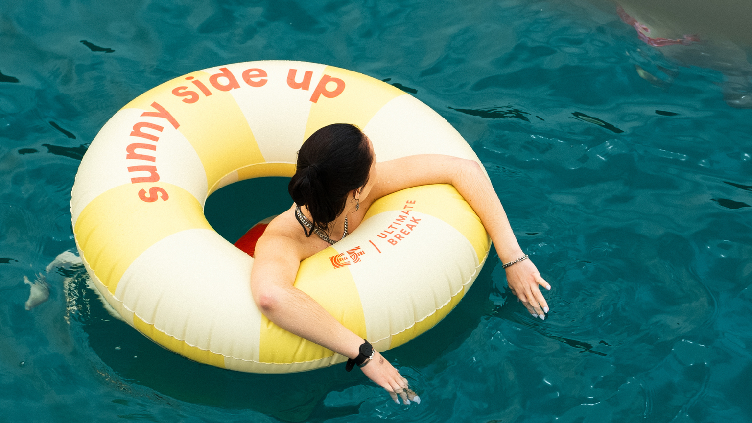 Person in a yellow and white inflatable ring labeled "sunny side up," floating in clear blue water.