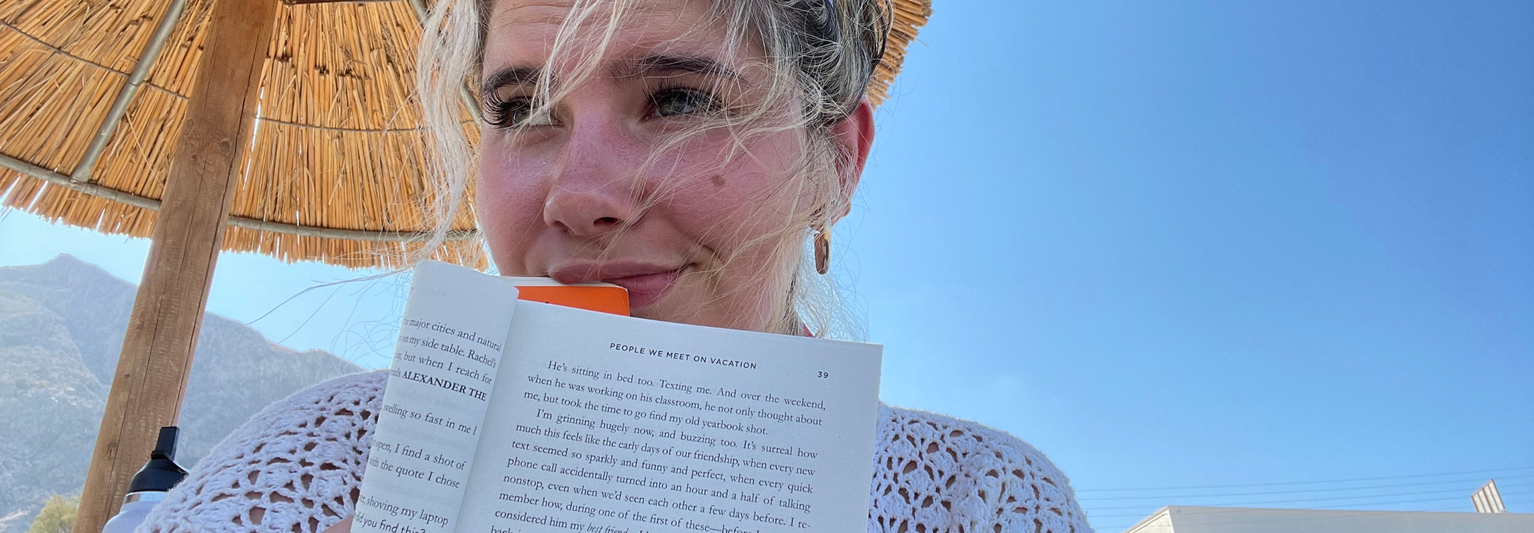 Woman under a straw umbrella, holding a book with windblown hair, against a clear blue sky and mountains in the background.