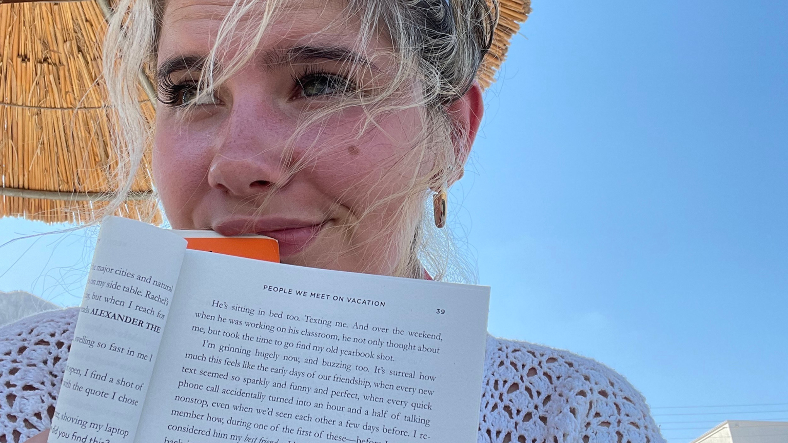 Woman under a straw umbrella, holding a book with windblown hair, against a clear blue sky and mountains in the background.