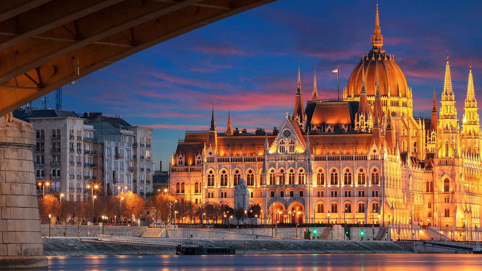 The illuminated Hungarian Parliament Building at dusk, viewed from under a bridge with a calm river reflecting the warm lights.
