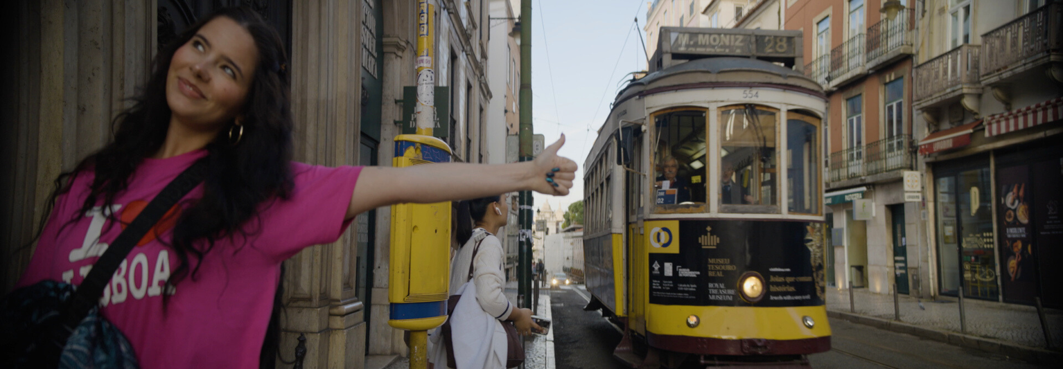 Woman in pink shirt gestures thumbs-up near a yellow tram on a narrow Lisbon street, with historic buildings in the background.