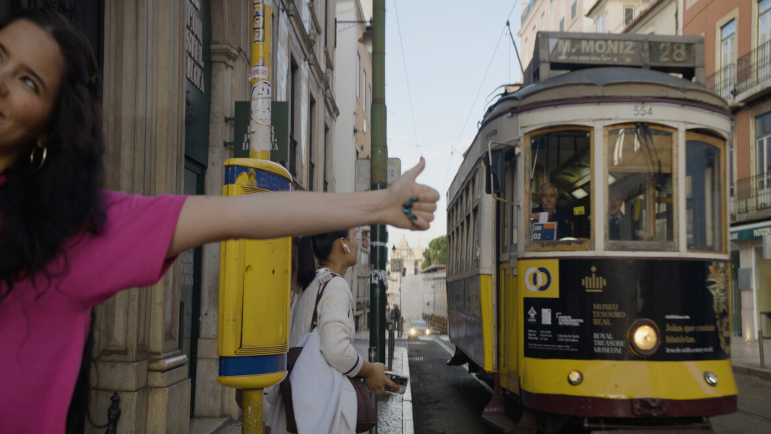 Woman in pink shirt gestures thumbs-up near a yellow tram on a narrow Lisbon street, with historic buildings in the background.