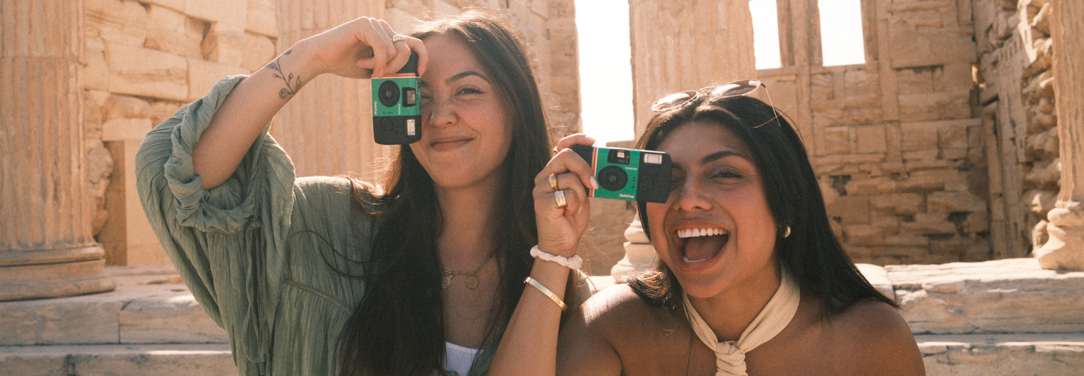 Two women holding up disposable cameras and standing in front of the Parthenon in Athens, Greece.
