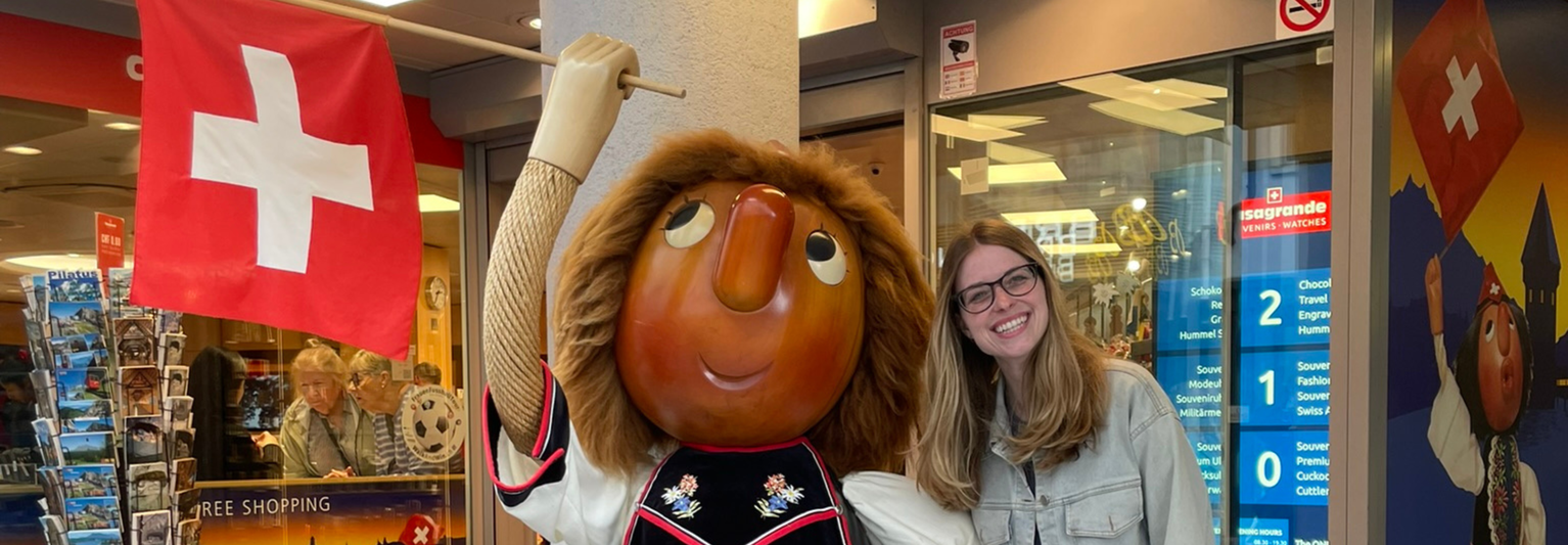 Woman smiling next to a large Swiss-themed mascot statue holding a flag. Storefront with souvenirs and Swiss flag visible in the background.