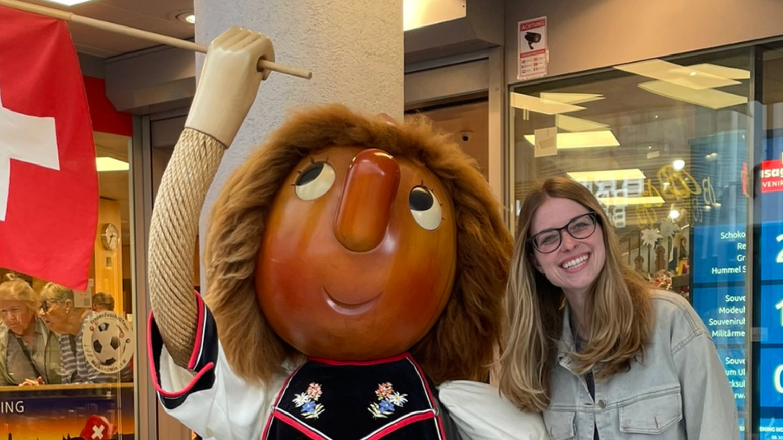 Woman smiling next to a large Swiss-themed mascot statue holding a flag. Storefront with souvenirs and Swiss flag visible in the background.