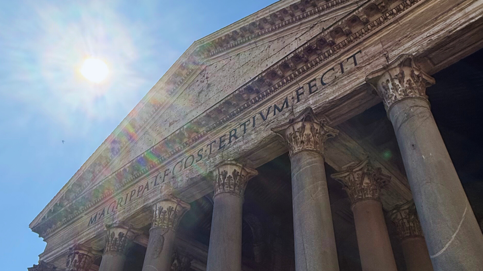 Sunlit view of the Pantheon in Rome, showcasing its grand columns and detailed pediment against a clear blue sky.