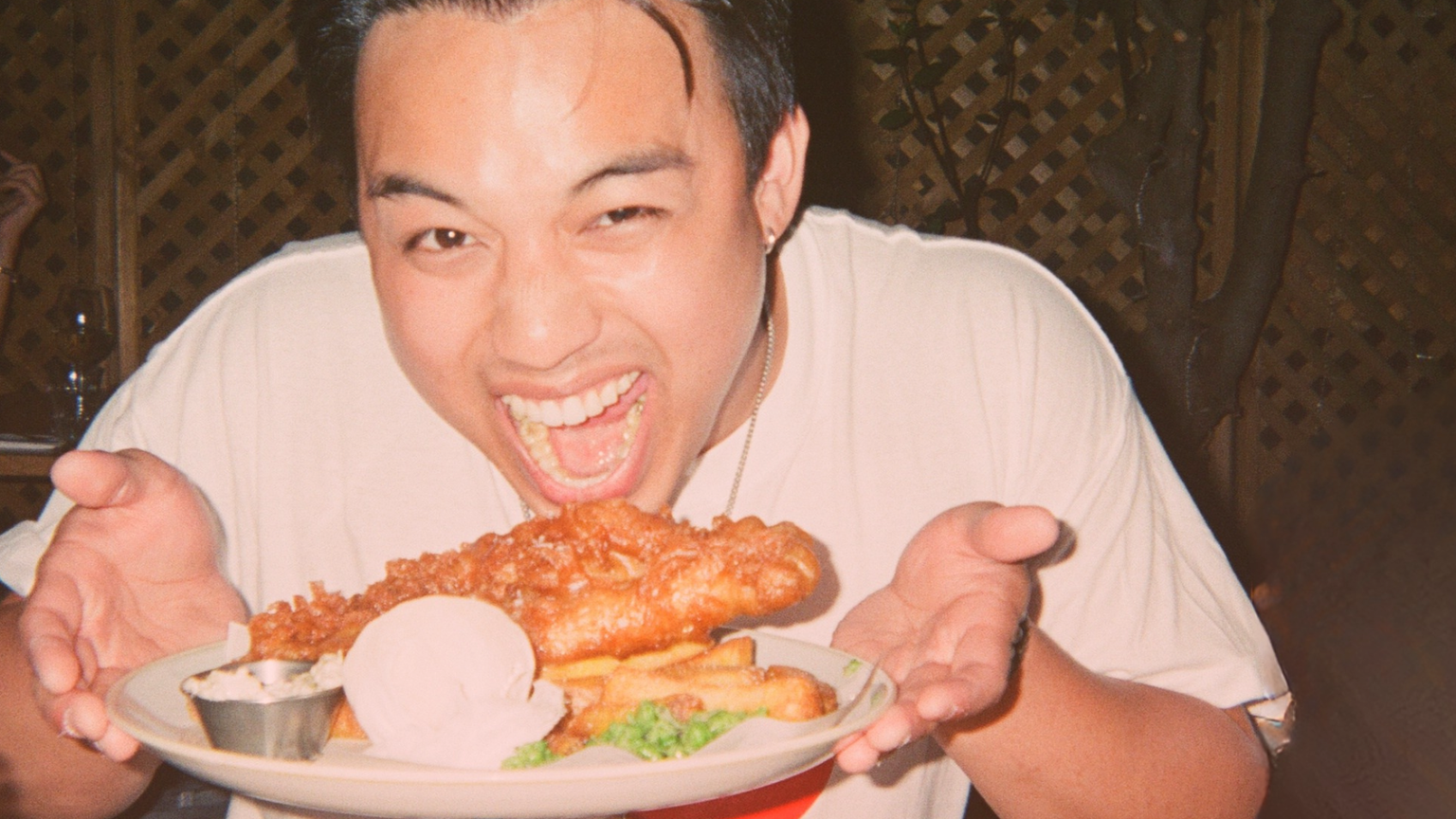 Person excitedly holding a plate of fish and chips, with a joyful expression, sitting at a table in a restaurant.