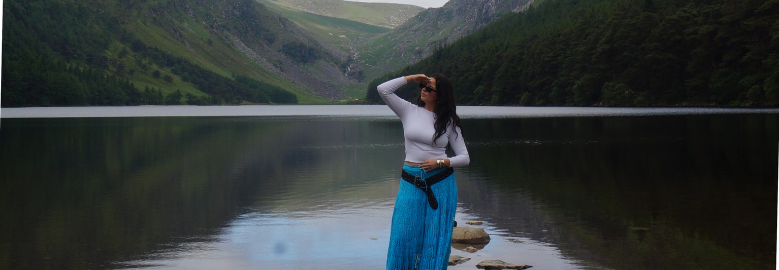 A woman in a white top and blue skirt stands on rocks by a calm lake, surrounded by lush green hills.