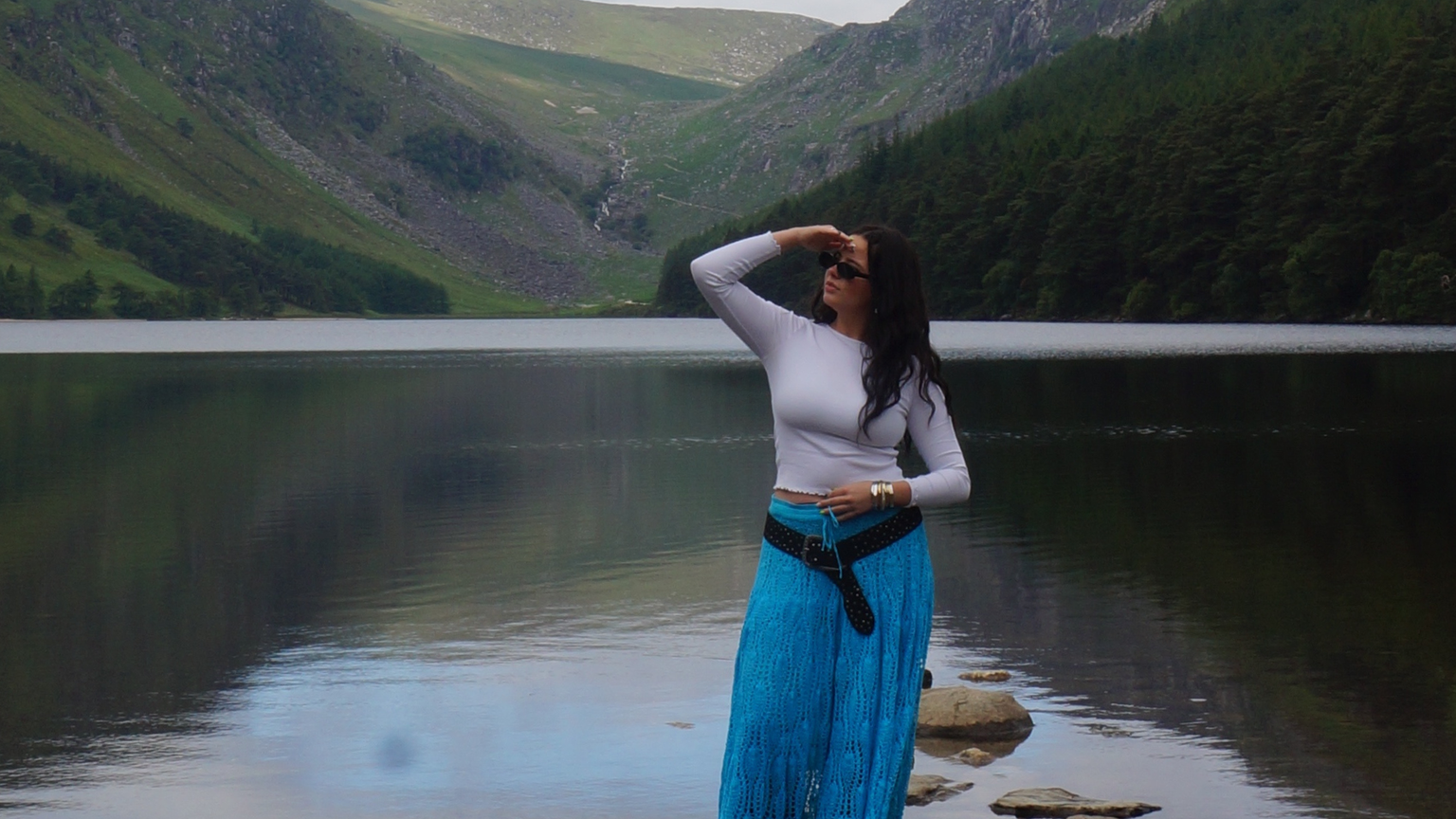 A woman in a white top and blue skirt stands on rocks by a calm lake, surrounded by lush green hills.