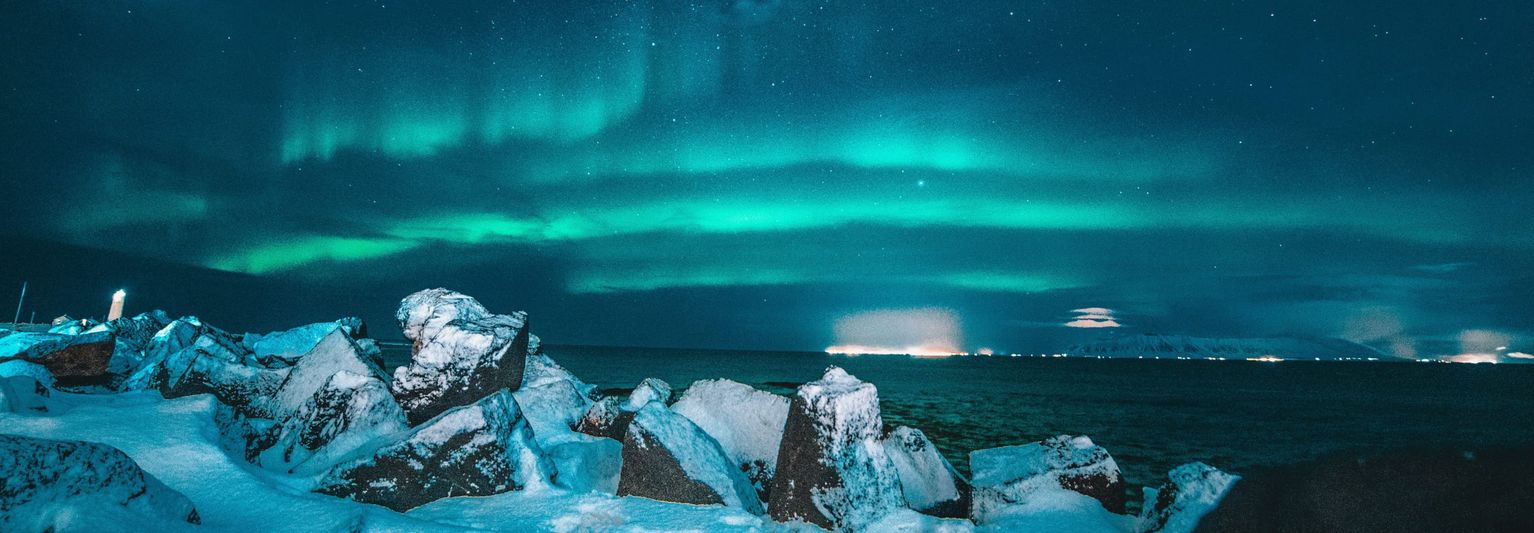 Northern lights illuminate the night sky over a rocky, snow-covered shoreline, with distant city lights along the horizon under a starry sky.