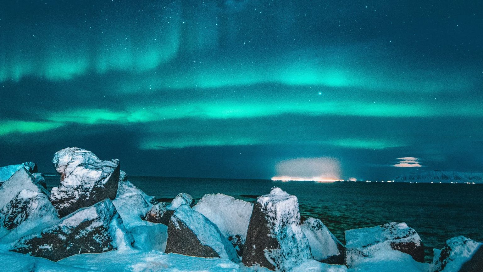 Northern lights illuminate the night sky over a rocky, snow-covered shoreline, with distant city lights along the horizon under a starry sky.