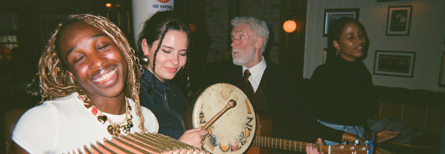 Four people enjoying music; one plays an accordion, another a drum, while the others sing and smile in a cozy, warmly lit room.