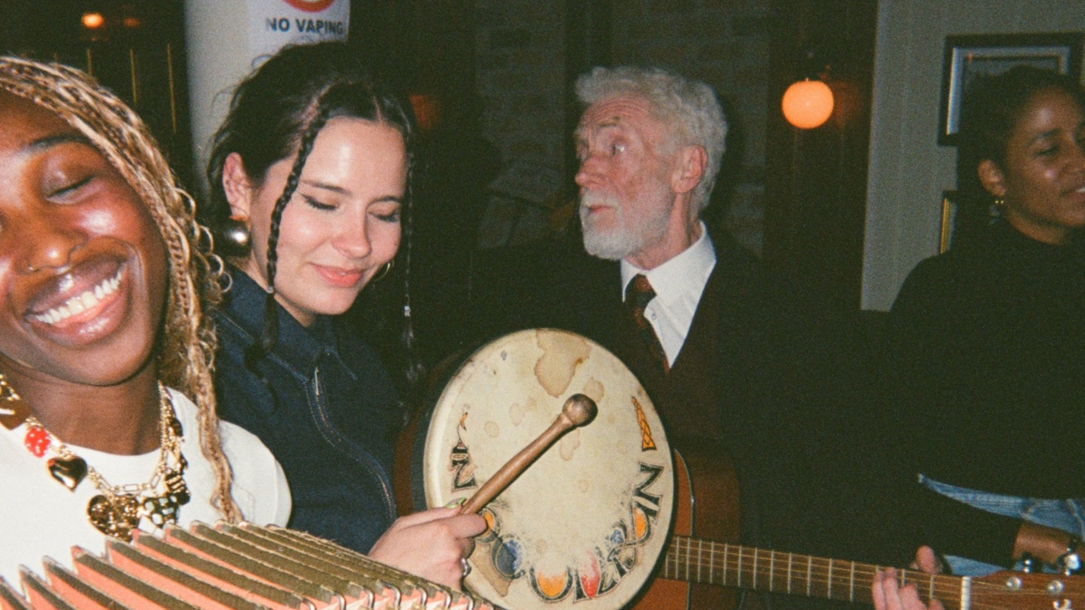 Four people enjoying music; one plays an accordion, another a drum, while the others sing and smile in a cozy, warmly lit room.