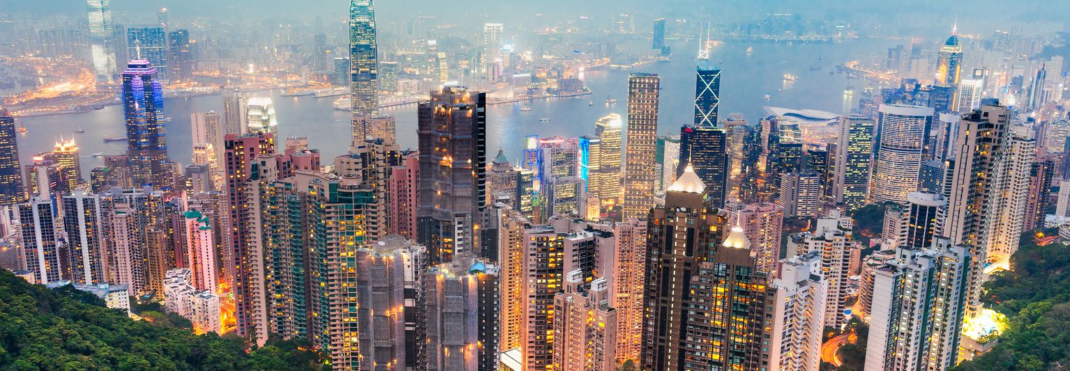 Aerial view of a vibrant cityscape with illuminated skyscrapers and a harbor in the background, surrounded by a misty sky.