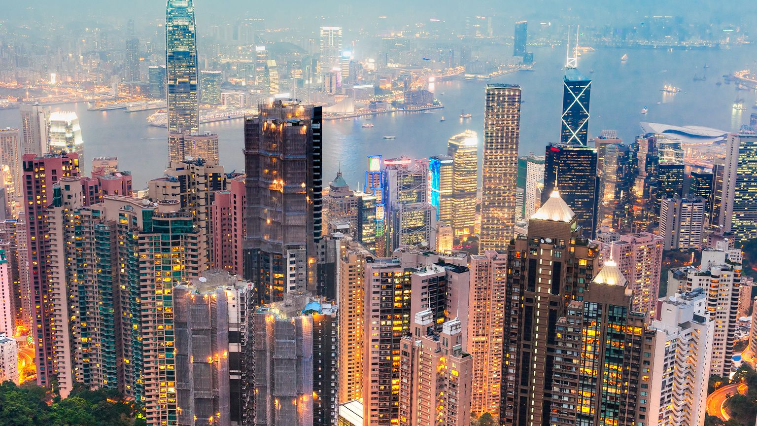 Aerial view of a vibrant cityscape with illuminated skyscrapers and a harbor in the background, surrounded by a misty sky.