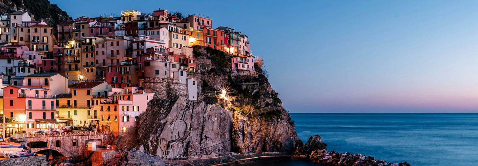 Colorful cliffside village with stacked houses overlooking a calm sea at dusk, illuminated by soft lights against a clear, fading sky.