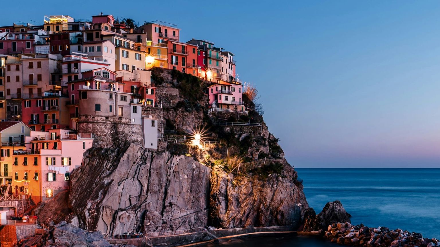 Colorful cliffside village with stacked houses overlooking a calm sea at dusk, illuminated by soft lights against a clear, fading sky.