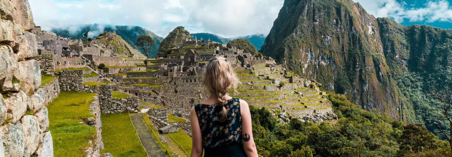 A person with blonde hair stands facing the ancient ruins of Machu Picchu, surrounded by green terraces and dramatic mountain peaks.