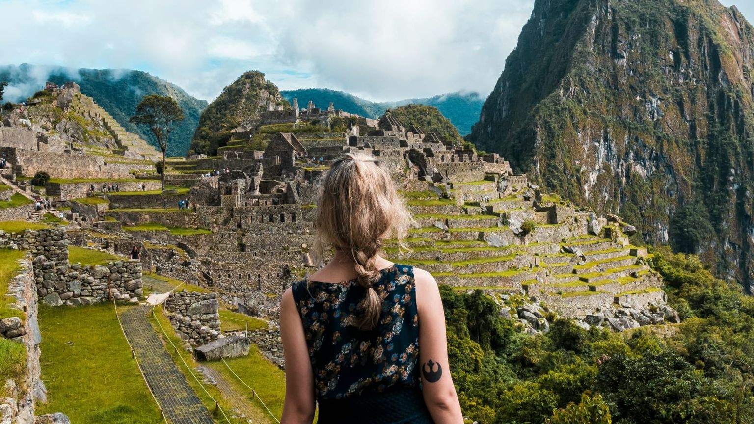 A person with blonde hair stands facing the ancient ruins of Machu Picchu, surrounded by green terraces and dramatic mountain peaks.