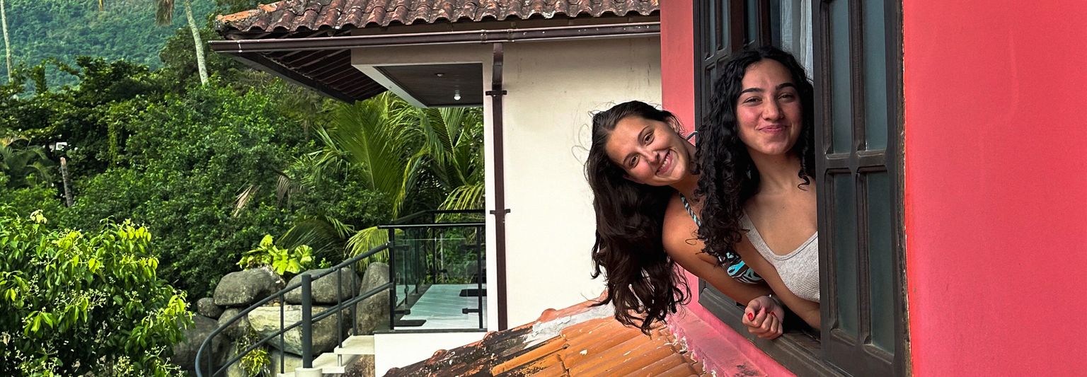 Two smiling women lean out of a red building's window, surrounded by lush greenery and a rooftop with tropical plants in the background.