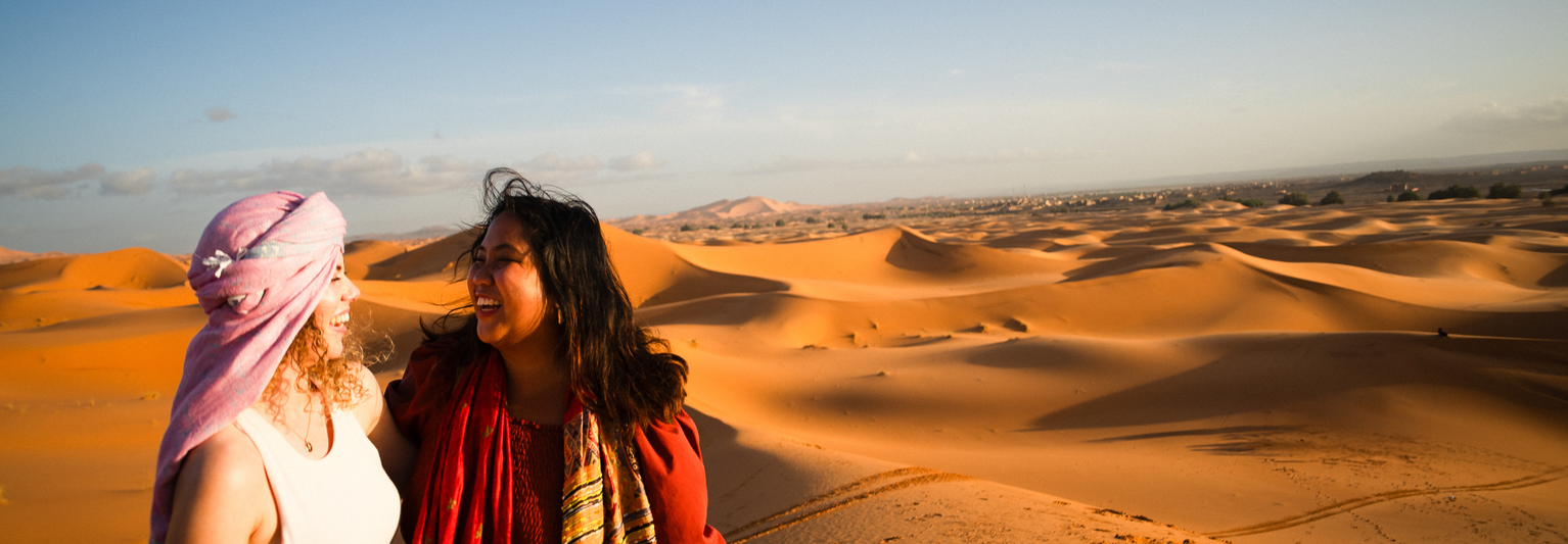 Two people smiling at each other in a desert landscape with vast sand dunes under a clear blue sky.