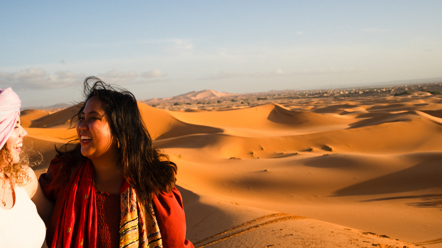 Two people smiling at each other in a desert landscape with vast sand dunes under a clear blue sky.