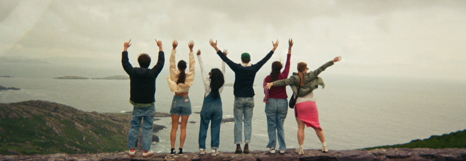 Six people stand on a cliff with arms raised, facing the ocean and cloudy sky, surrounded by green hills.