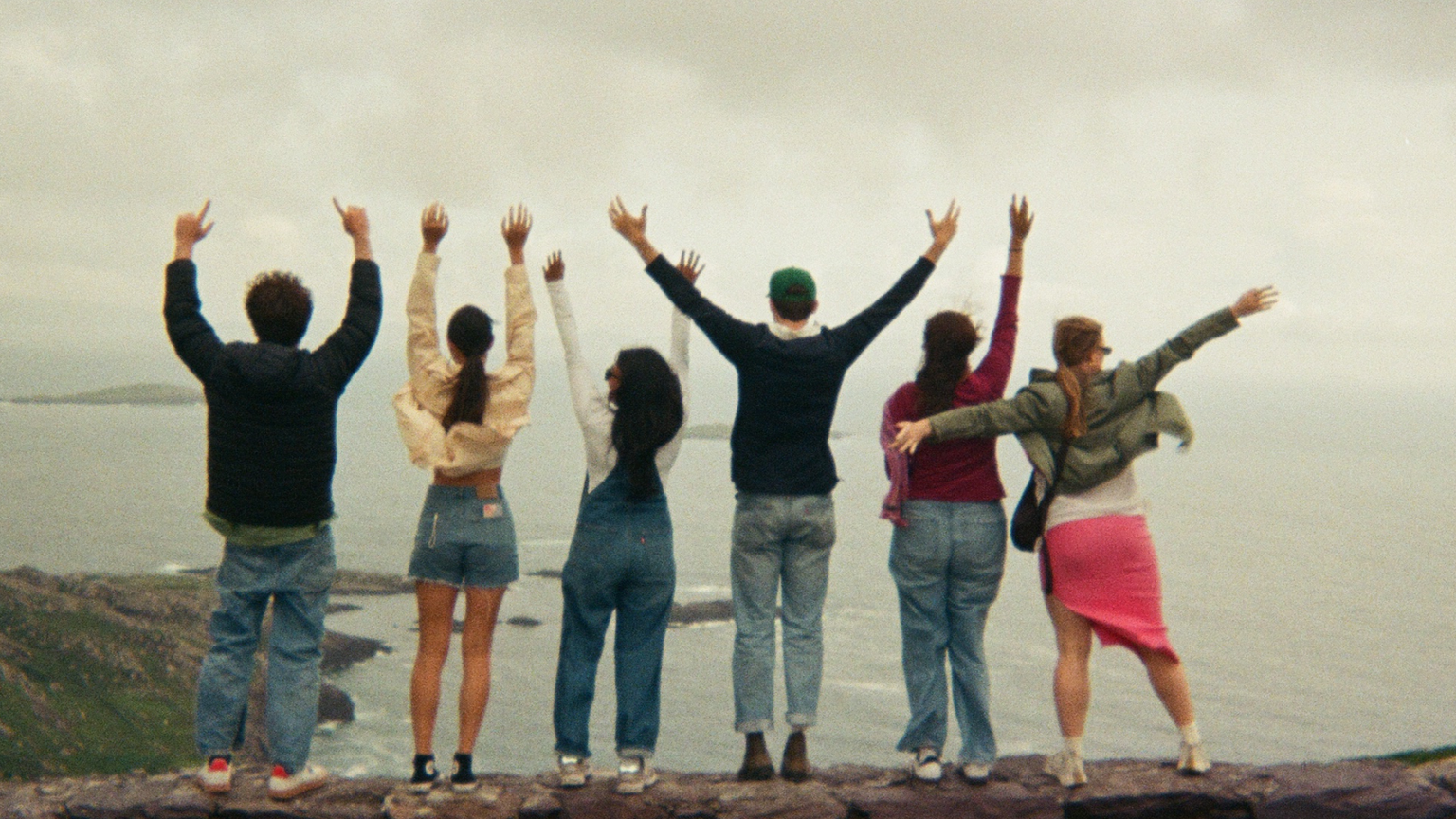 Six people stand on a cliff with arms raised, facing the ocean and cloudy sky, surrounded by green hills.