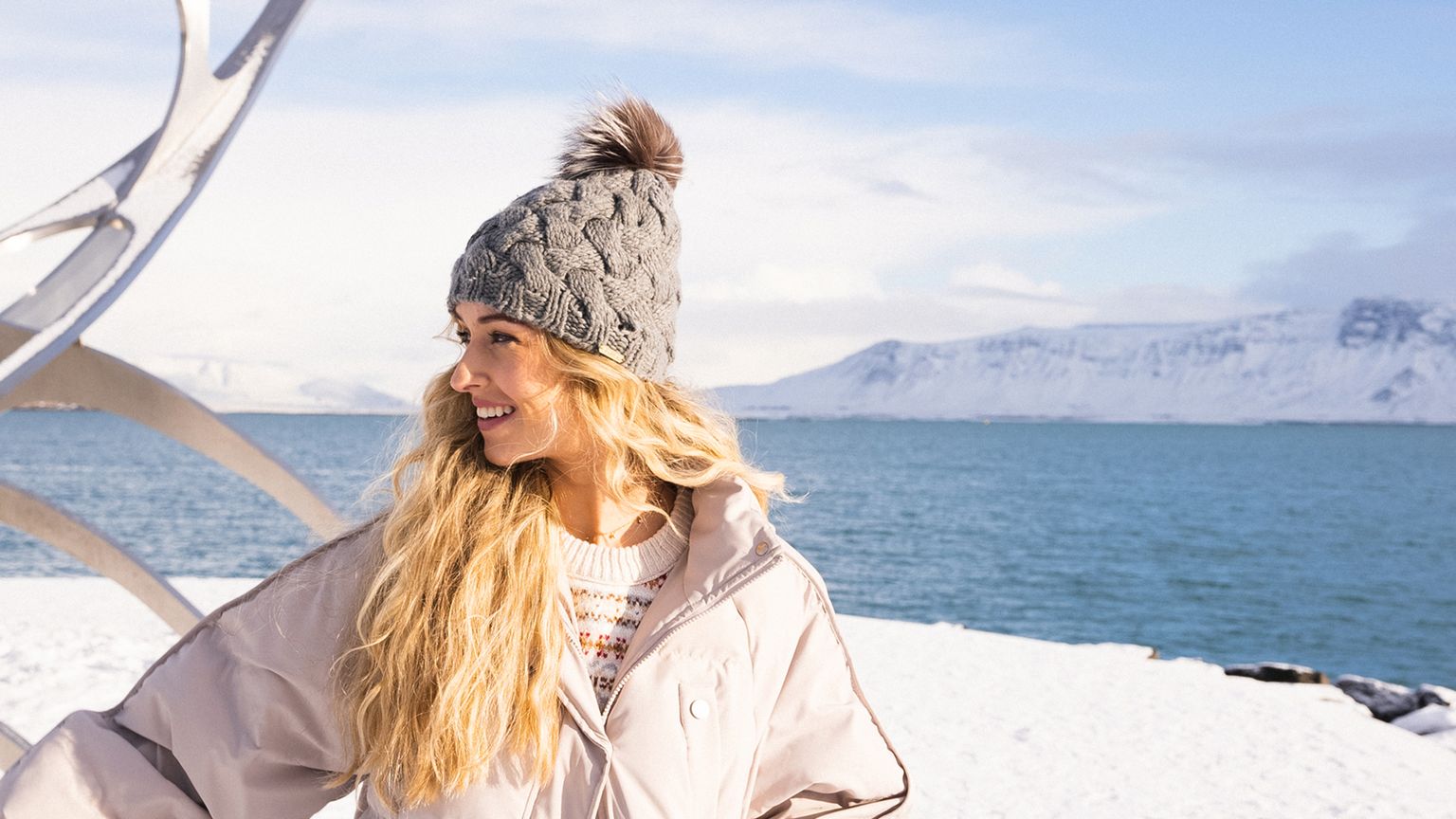 A woman in a winter coat and knitted hat smiles by the sea, with mountains and a metallic sculpture in the background on a snowy day.