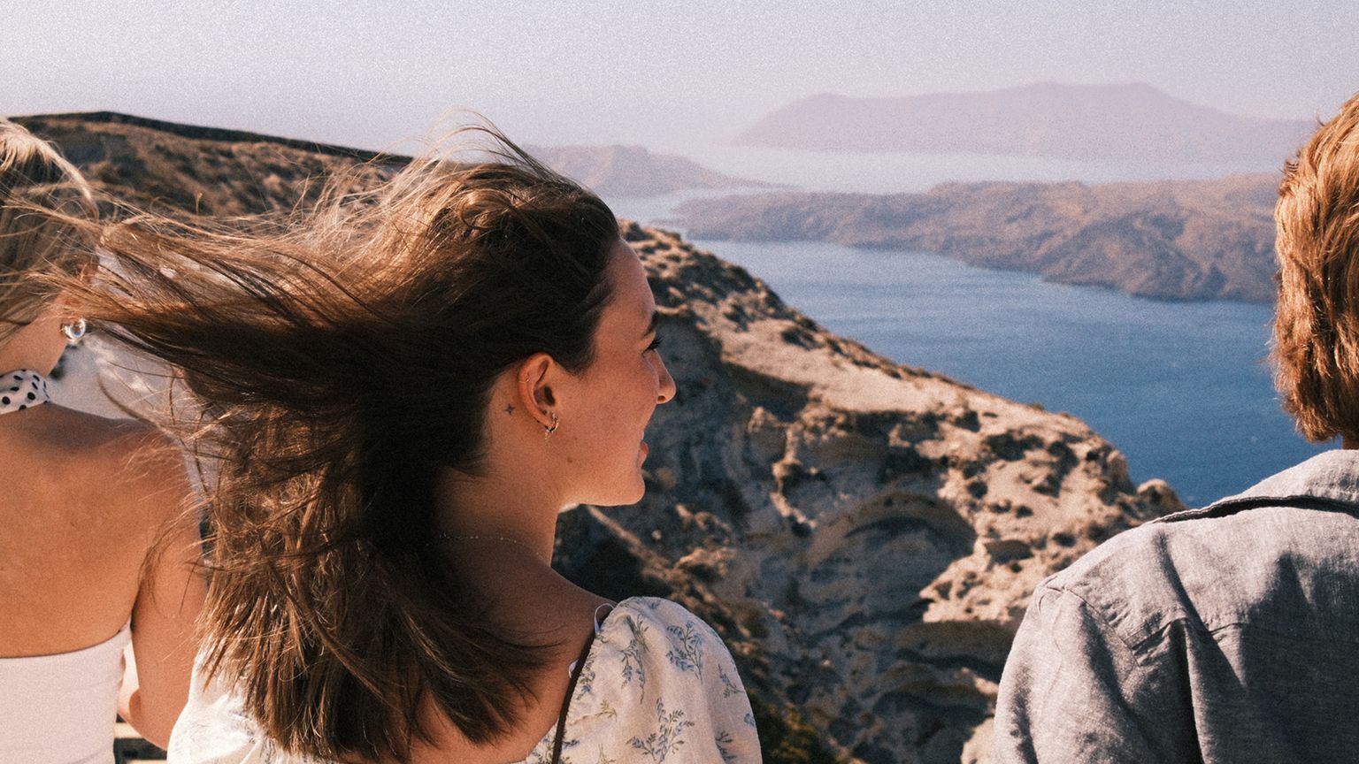 Three people enjoying a scenic view of cliffs and the sea on a sunny day, with hair blowing in the wind.
