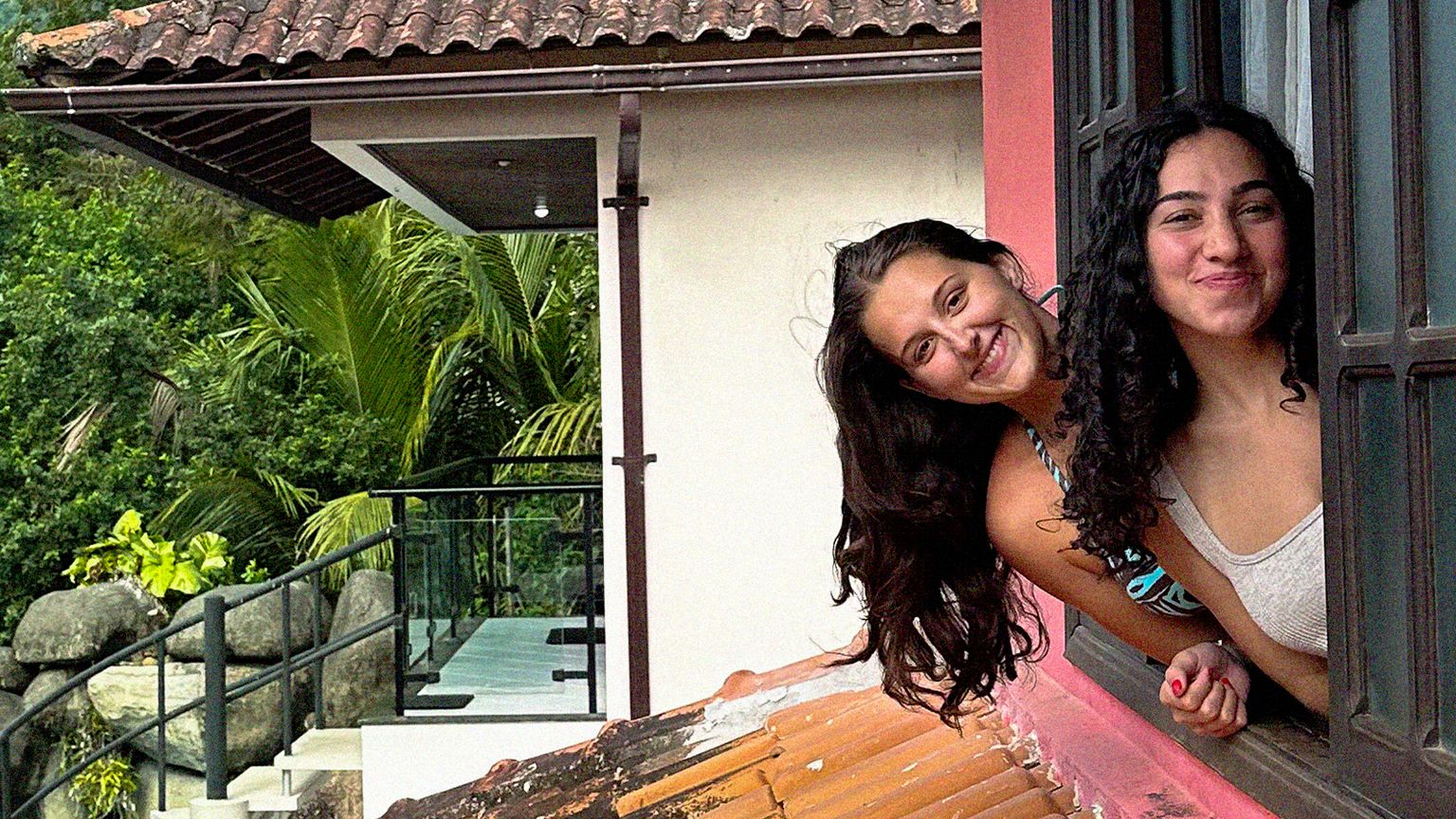 Two women smiling and leaning out of a window in a red building, surrounded by lush greenery and trees.