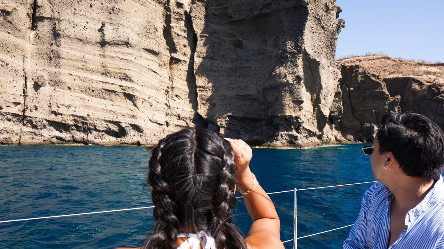 Two people on a boat admire tall, rocky cliffs with deep blue water beneath a clear sky.